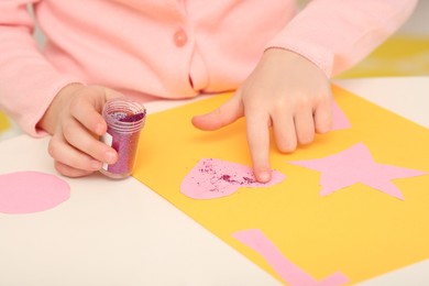 Photo of Girl using colorful glitter while making paper card at desk in room, closeup. Home workplace