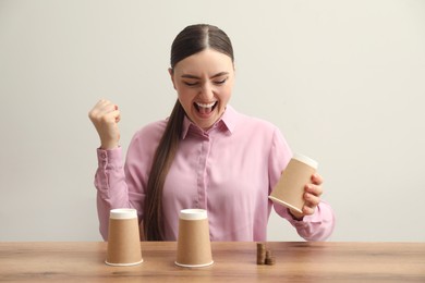 Photo of Shell game. Emotional woman showing stack of coins under cup at wooden table