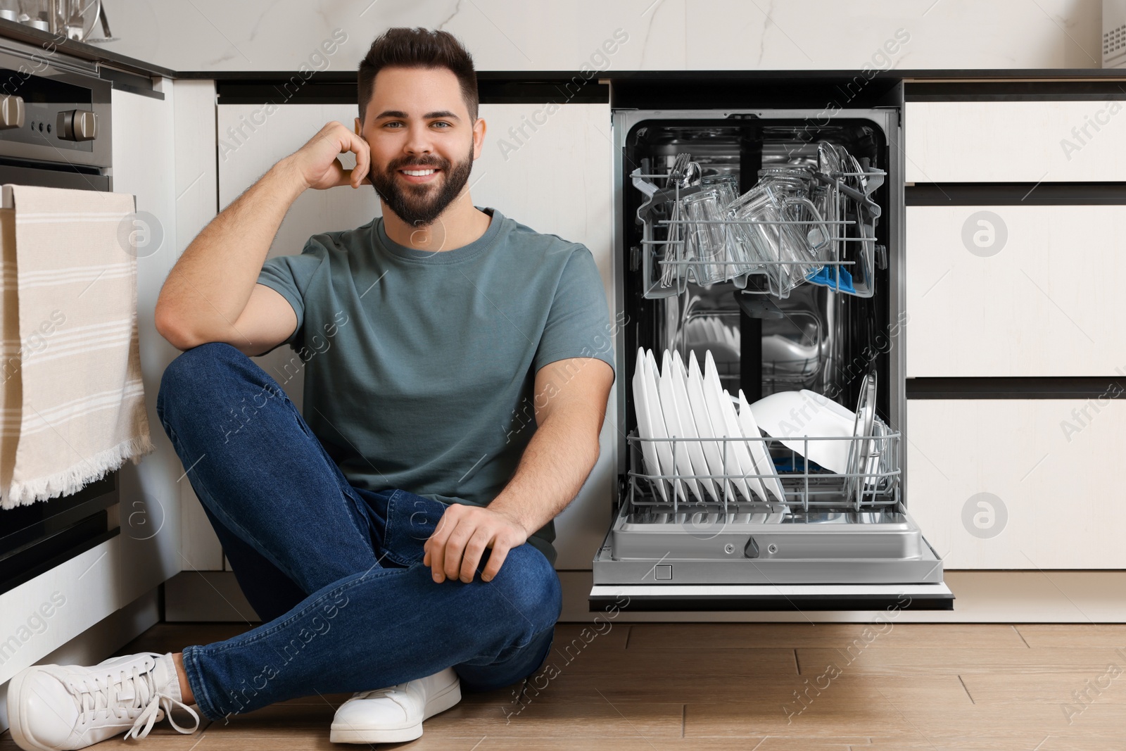 Photo of Smiling man sitting near open dishwasher in kitchen