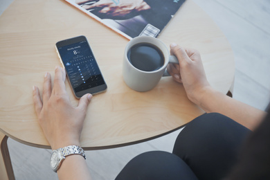 Woman with cup of coffee using weather forecast app on smartphone indoors, closeup