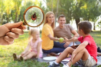 Family having picnic in park and don't even suspect about hidden danger in green grass. Woman showing tick with magnifying glass, selective focus