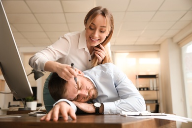 Young woman drawing on colleague's face while he sleeping in office. Funny joke