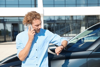 Photo of Young man checking time while talking on phone near modern car, outdoors
