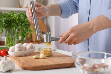 Woman squeezing garlic with press at white wooden table in kitchen, closeup