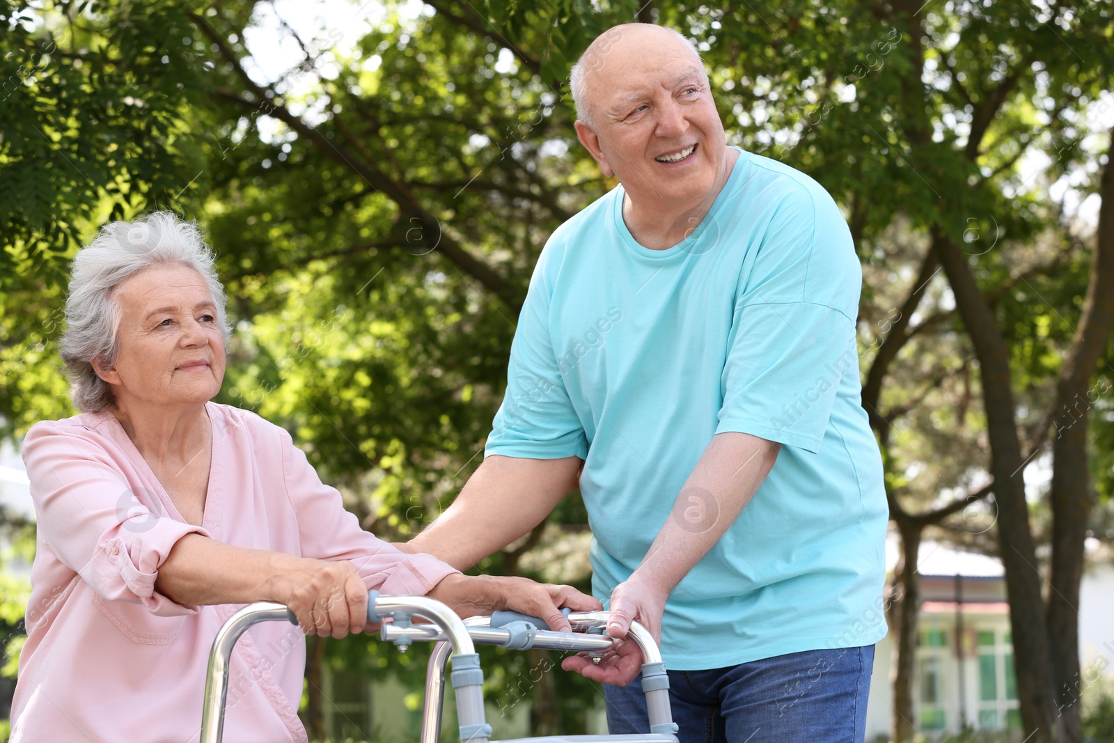 Photo of Elderly man helping his wife with walking frame outdoors