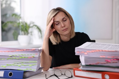 Overwhelmed woman sitting at table with stacks of documents and folders in office