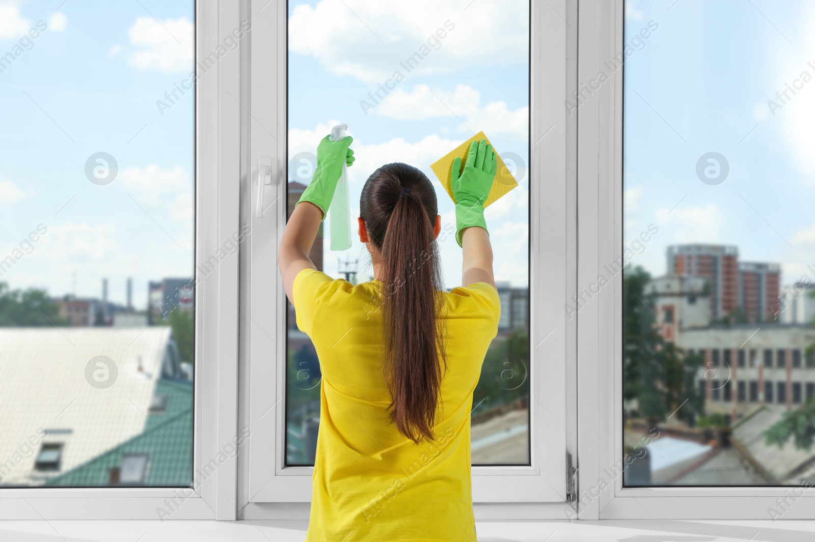 Photo of Woman cleaning window glass with sponge cloth and spray indoors, back view