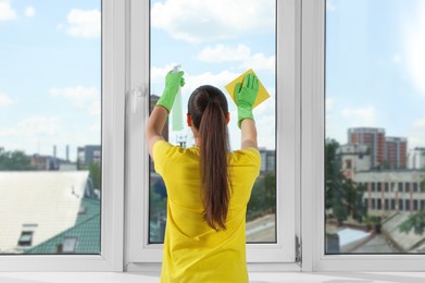 Photo of Woman cleaning window glass with sponge cloth and spray indoors, back view