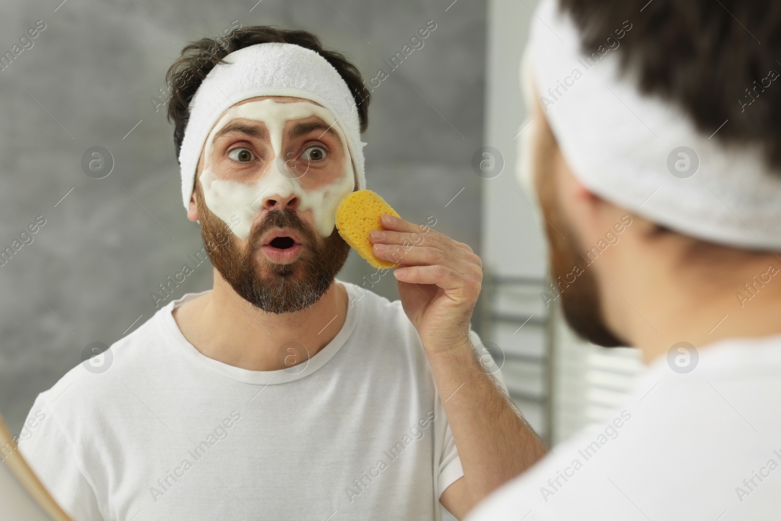 Photo of Emotional man with headband washing his face using sponge near mirror in bathroom