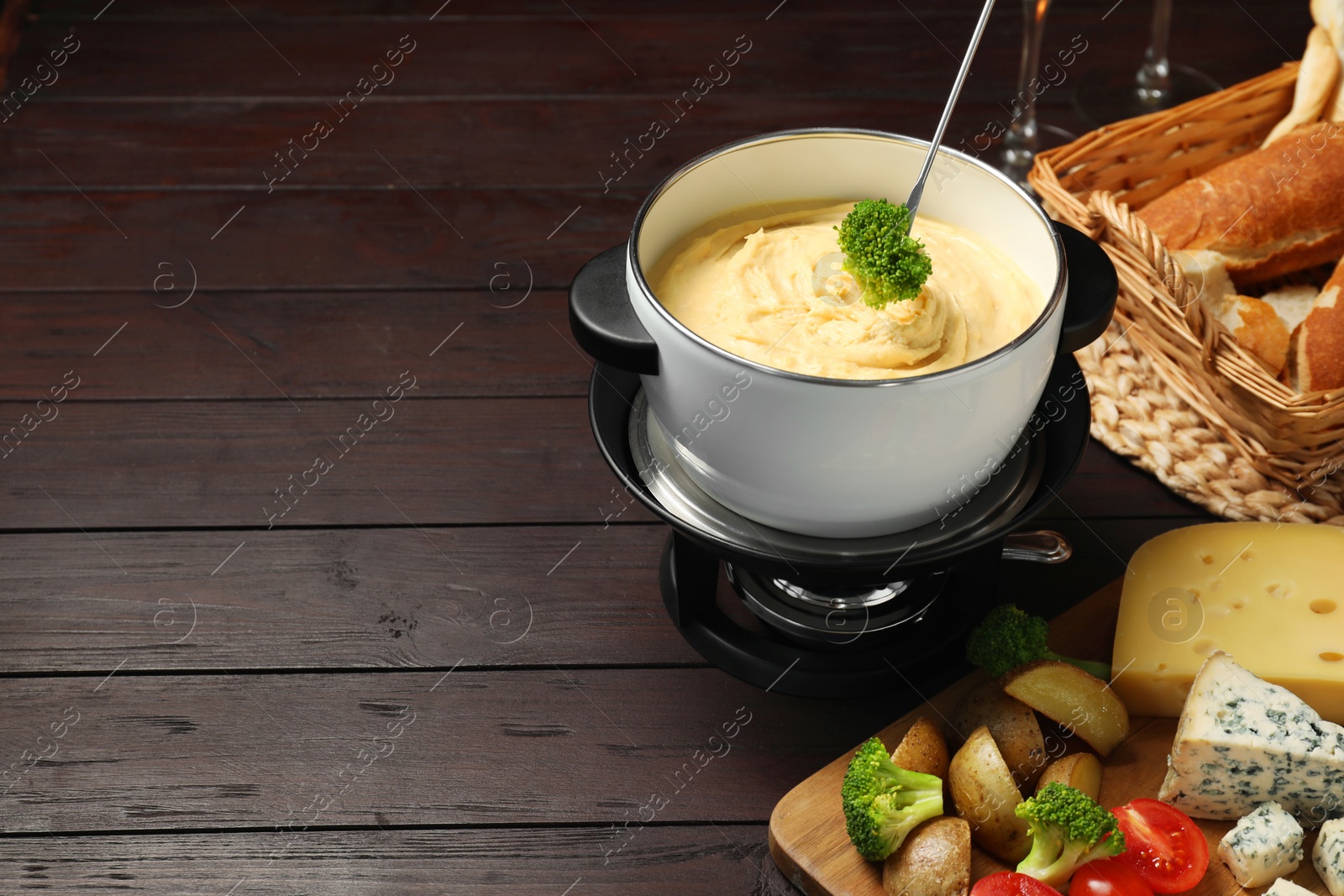 Photo of Dipping piece of broccoli into fondue pot with melted cheese at wooden table with snacks, closeup. Space for text