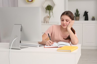 Photo of Tired girl writing in notepad while using computer at desk in room. Home workplace