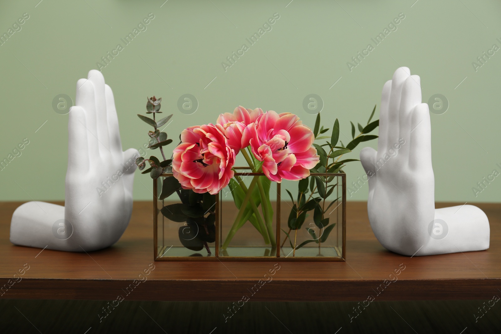 Photo of Glass holder with green branches and beautiful flowers near hand shaped bookends on wooden table indoors. Stylish interior design
