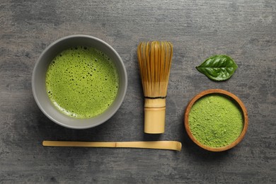 Photo of Cup of fresh matcha tea, bamboo whisk, spoon and green powder on dark grey table, flat lay