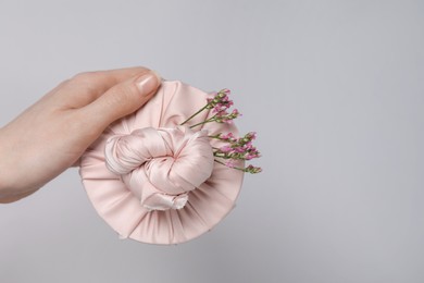 Photo of Furoshiki technique. Woman holding gift packed in fabric and beautiful pink flowers on gray background, closeup with space for text