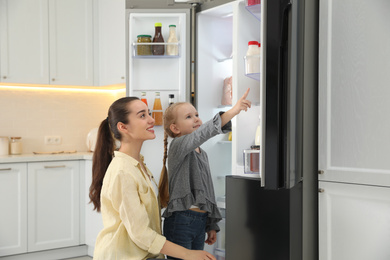 Young mother with daughter near open refrigerator in kitchen