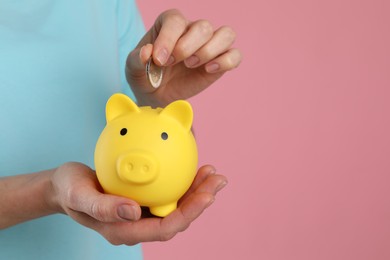 Woman putting coin into yellow piggy bank on pink background, closeup. Space for text