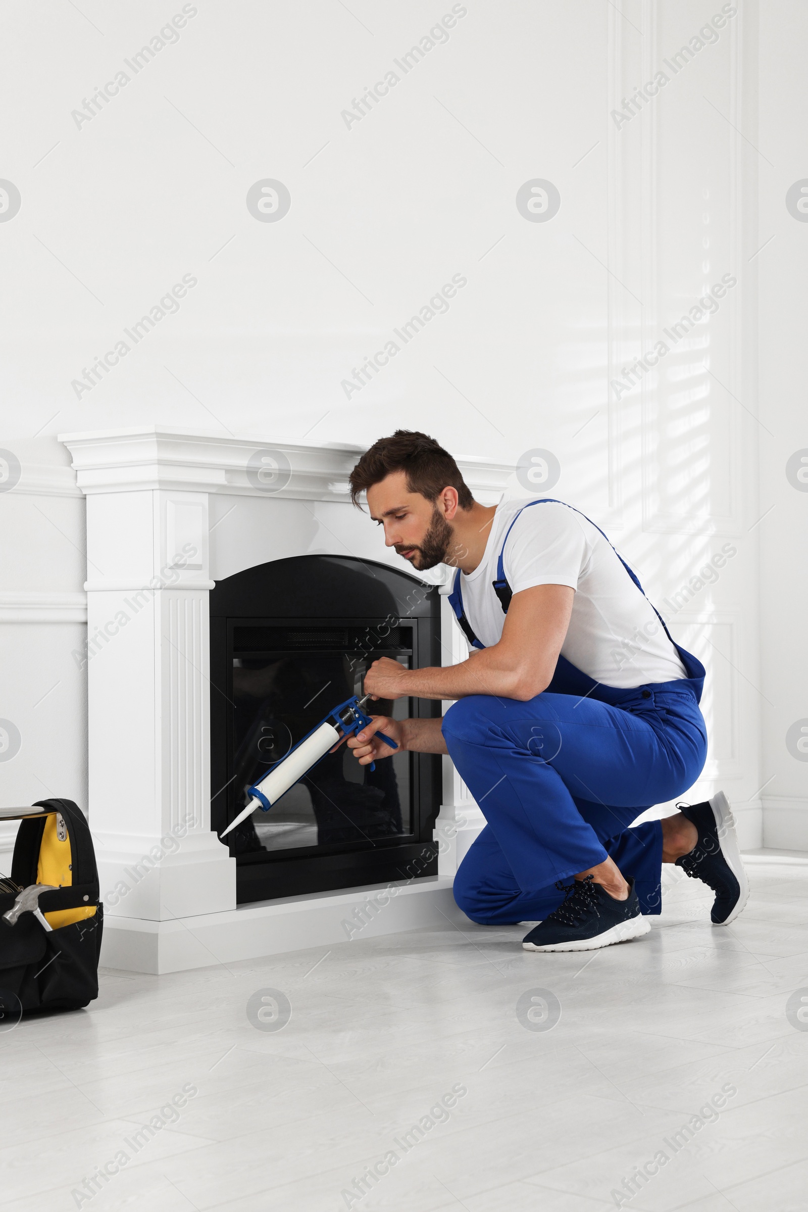 Photo of Professional technician sealing electric fireplace with caulk near white wall in room