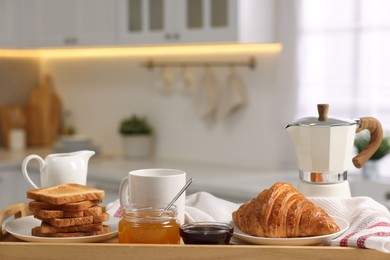 Photo of Breakfast served in kitchen. Toasts, honey, jam, fresh croissant, coffee and pitcher of milk in wooden tray, closeup