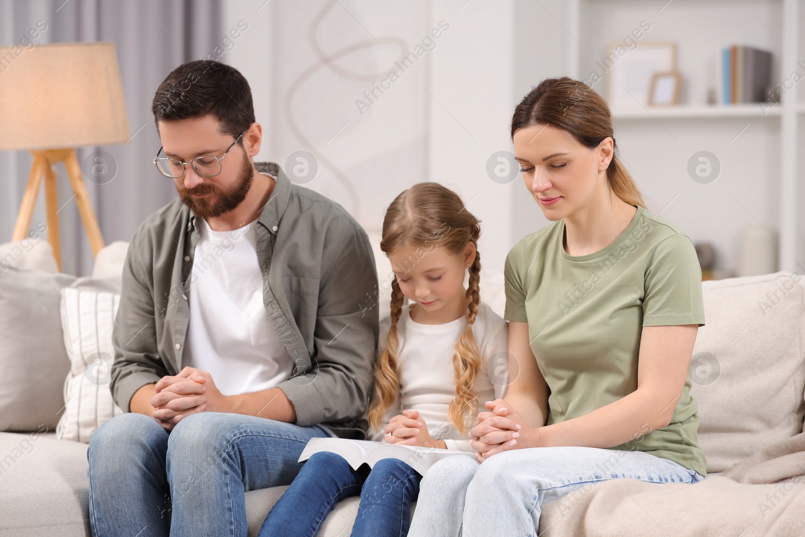 Photo of Girl and her godparents praying together on sofa at home