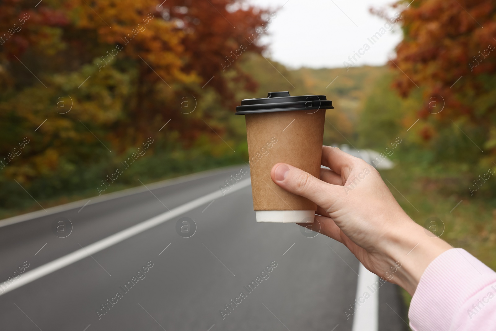 Photo of Woman holding takeaway cardboard coffee cup near road outdoors, closeup. Autumn season