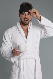 Young man in bathrobe with cup of coffee on grey background