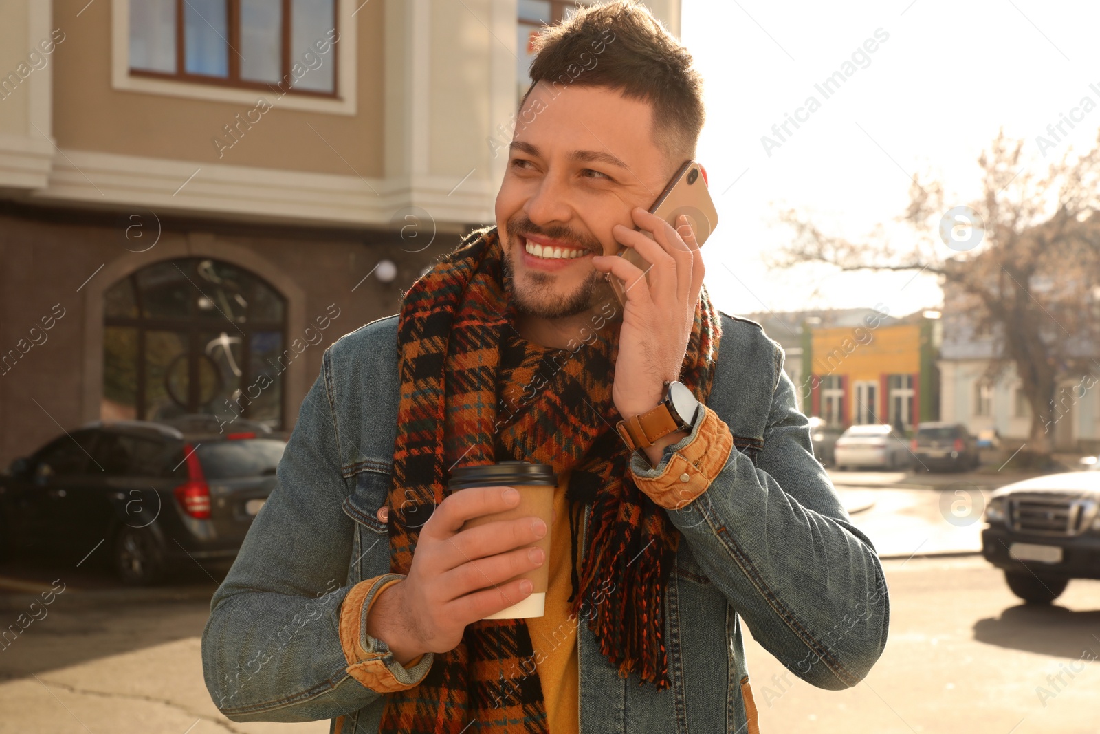 Photo of Man with cup of coffee talking on smartphone on city street in morning