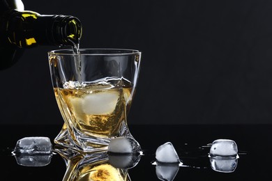 Photo of Pouring tasty whiskey from bottle into glass with ice at mirror table against black background, closeup. Space for text