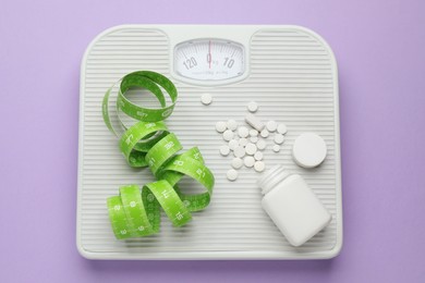 Photo of Scales with weight loss pills and measuring tape on violet background, top view