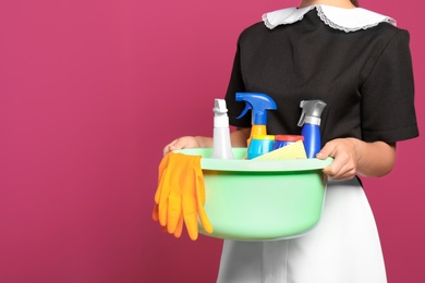 Young chambermaid holding plastic basin with detergents on color background