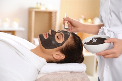 Cosmetologist applying black mask onto woman's face in spa salon