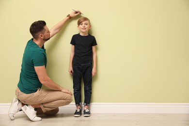 Father measuring height of his son near light wall indoors. Space for text