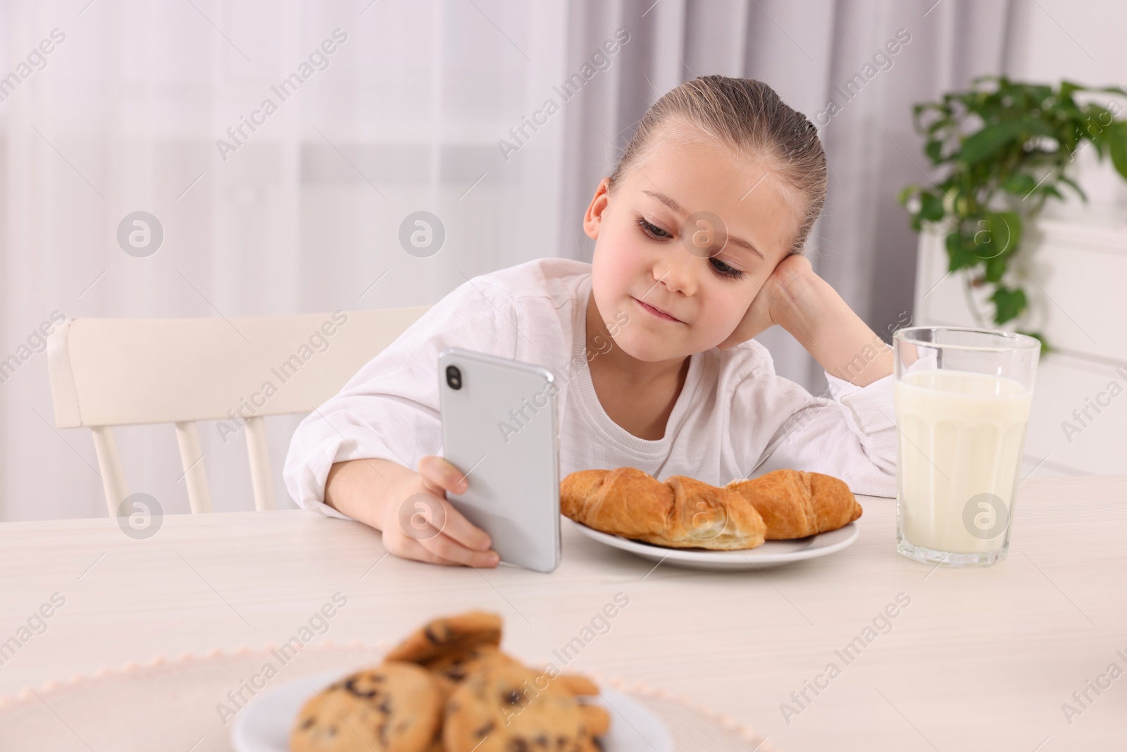 Photo of Little girl using smartphone while having breakfast at table indoors. Internet addiction