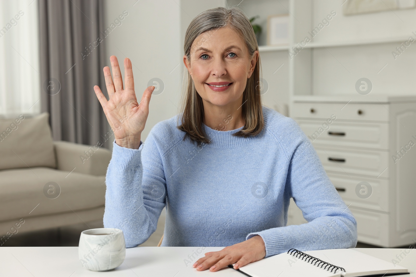 Photo of Happy woman waving hello at white table indoors