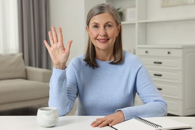 Happy woman waving hello at white table indoors