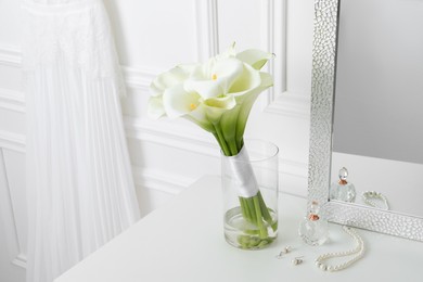 Beautiful calla lily flowers tied with ribbon in glass vase, bottle of perfume and jewelry on white table