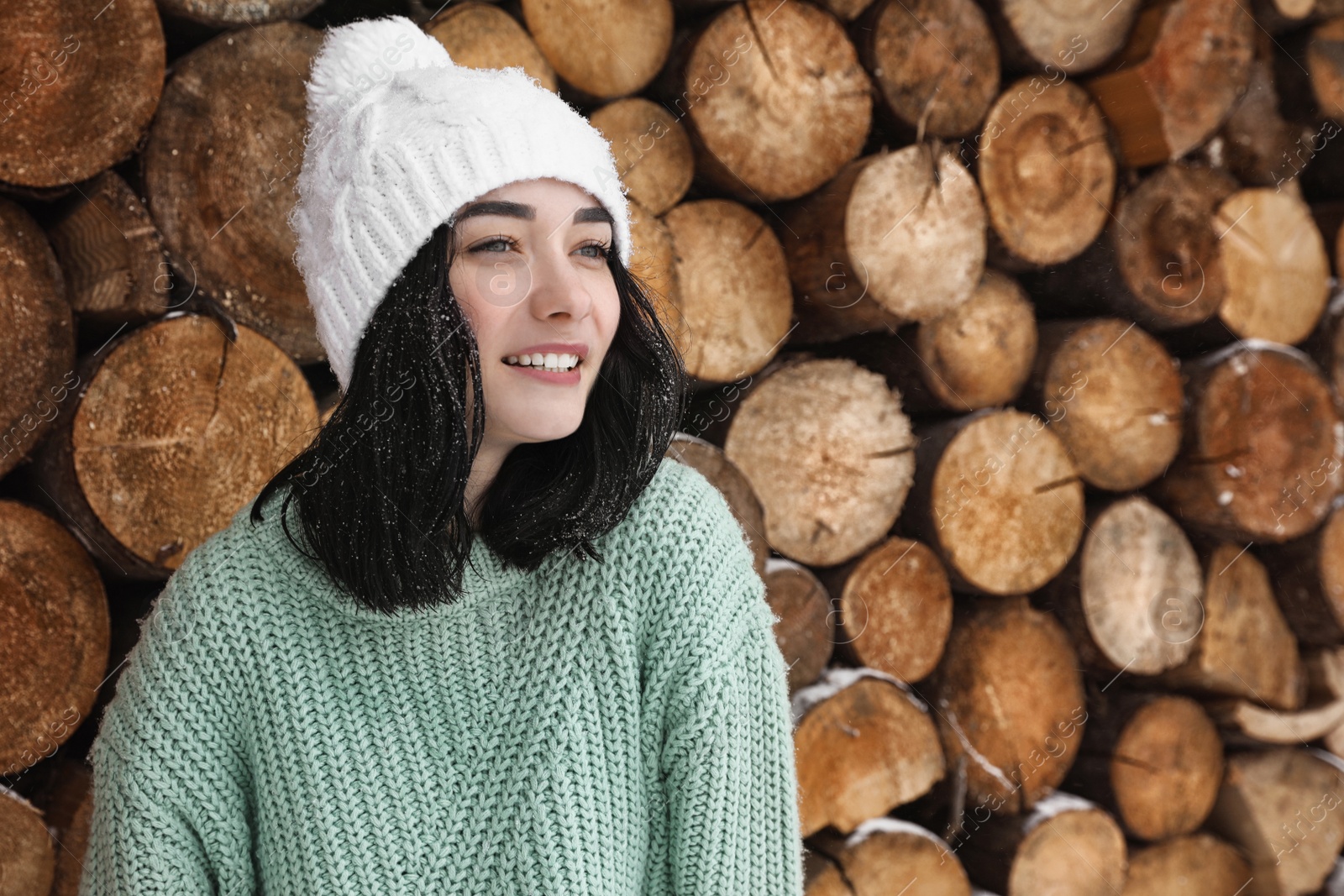 Photo of Young woman wearing warm sweater and hat near stack of firewood. Winter season