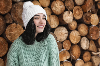 Young woman wearing warm sweater and hat near stack of firewood. Winter season