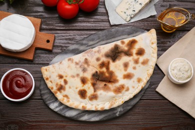Photo of Delicious cheese calzone, sauces and ingredients on wooden table, flat lay