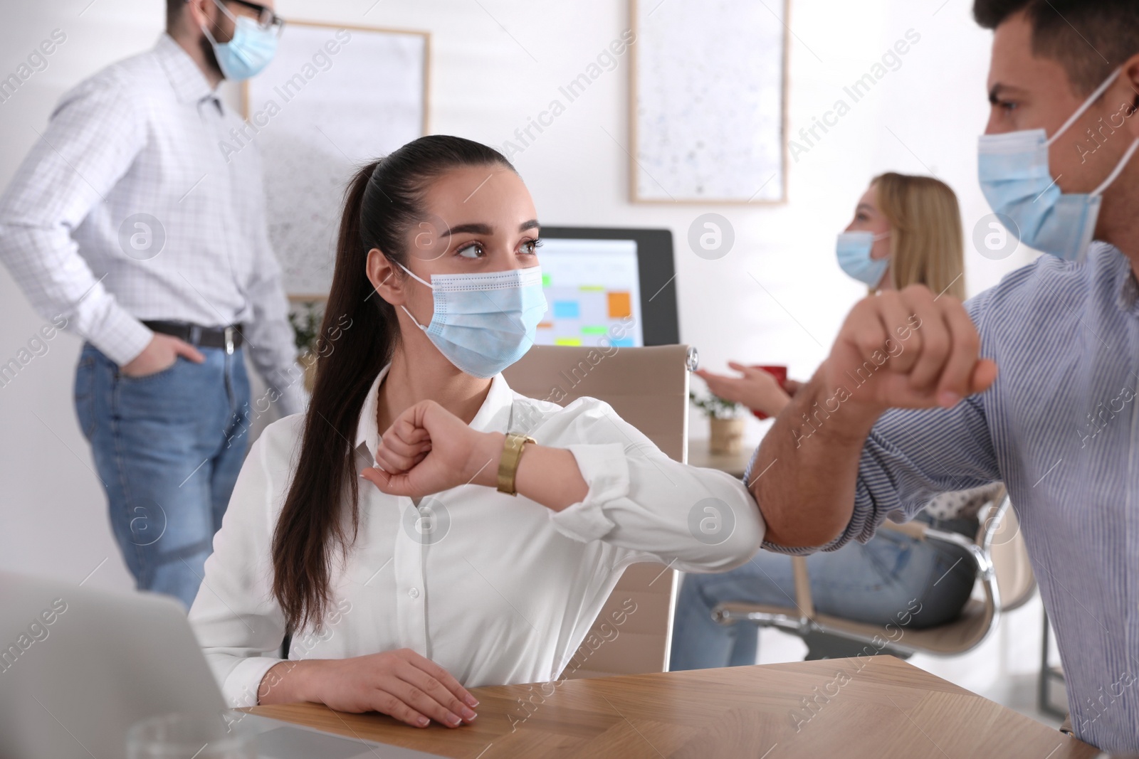 Photo of Coworkers with protective masks making elbow bump in office. Informal greeting during COVID-19 pandemic