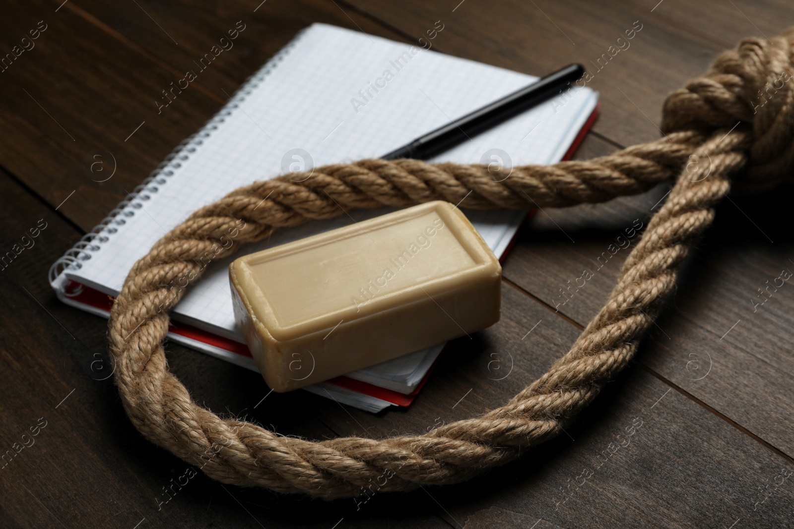 Photo of Rope noose, soap bar and blank notebook with pen on wooden table, closeup