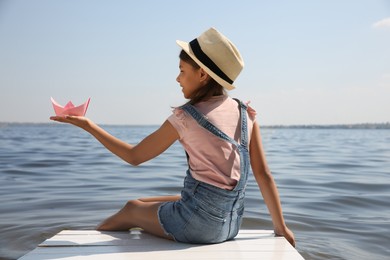 Cute little girl playing with paper boat on wooden pier near river