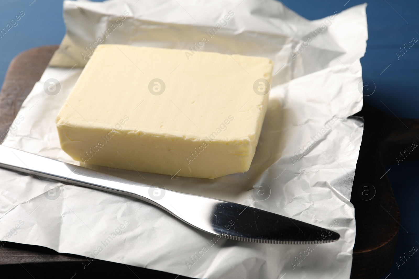 Photo of Block of tasty butter in open foil packaging and knife on blue wooden table, closeup