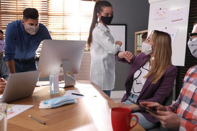 Photo of Coworkers with protective masks making elbow bump in office. Informal greeting during COVID-19 pandemic