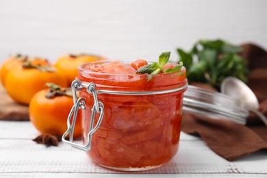 Photo of Jar of tasty persimmon jam and mint on white wooden table, closeup