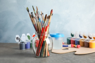 Photo of Glass jar with brushes and different paints on table against color background. Space for text