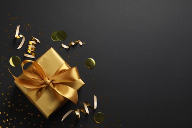 Golden gift box with confetti and streamers on black background, flat lay. Space for text