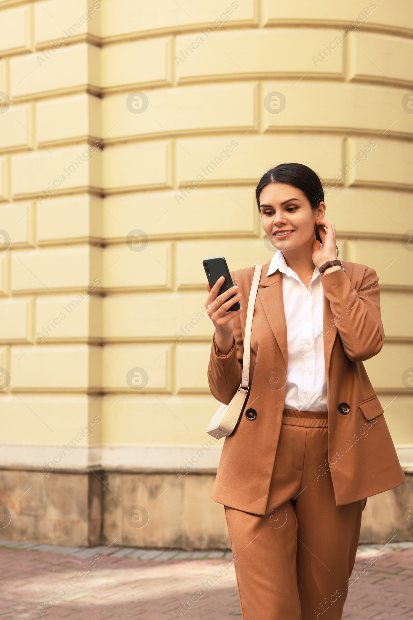 Photo of Young beautiful woman in stylish suit using smartphone near pale yellow building outdoors