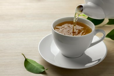 Photo of Pouring green tea into white cup with saucer and leaves on wooden table, closeup. Space for text