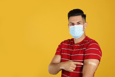 Photo of Vaccinated man with protective mask showing medical plaster on his arm against yellow background. Space for text