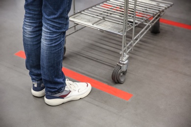Photo of Person with shopping cart standing behind taped floor marking in store for social distance, closeup. Preventive measure during coronavirus pandemic